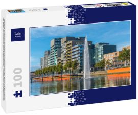 Lais Puzzle - Lelystad: Einheimische gehen und überqueren den Teich mit Brunnen in der Mitte mit modernen Geschäftsgebäuden und Häusern im Hintergrund - 100 Teile